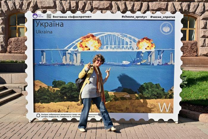 Sieviete reaģē, pozējot lielas pastmarkas priekšā, kurā attēlots sprādziens uz Krimas tilta, kas novietota Ukrainas galvaspilsētas Kijevas centrā 2022. gada 8. oktobrī. Foto: AFP/Scanpix