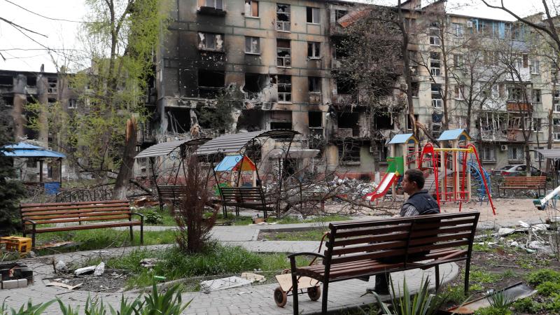 Vīrietis sēž uz soliņu pie iznīcinātas dzīvojamās ēkas Mariupolē
