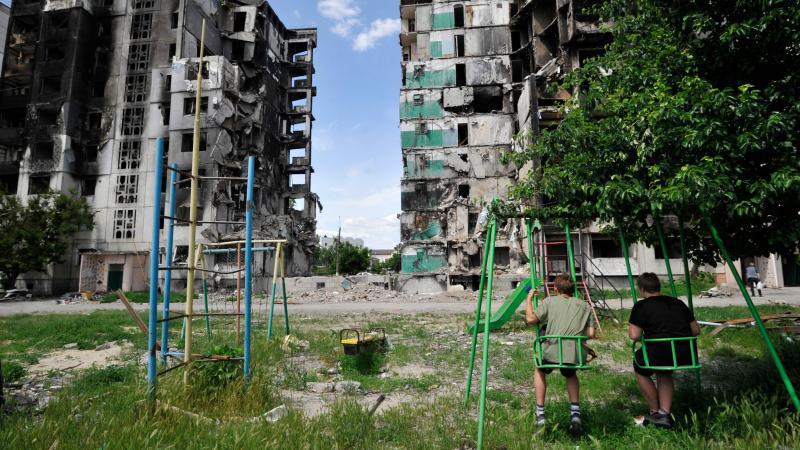 Divi zēni sēž šūpolēs, raugoties uz izpostītu dzīvojamo ēku Borodjankā